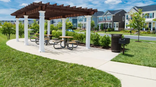 A pergola with a grill is located in a grassy park, across the street from single-family housing in a suburban style.