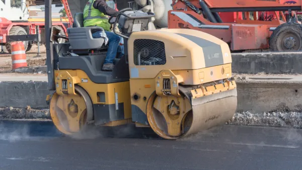 A man operates a yellow roller to smooth out fresh pavement on a road.
