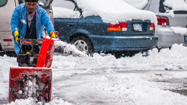 A man pushes a snow blower to clear the ground of an outdoor parking lot filled with cars.
