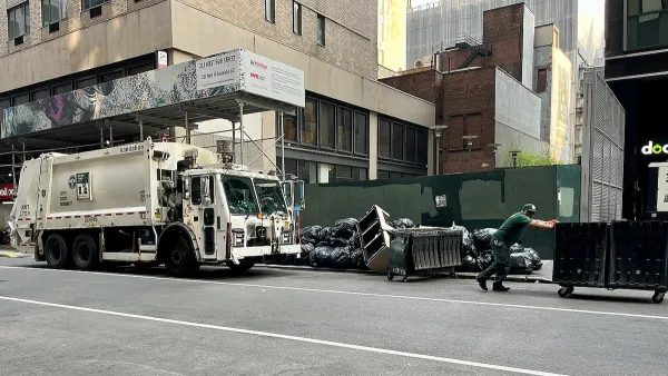 White waste management sanitation garbage truck and worker pushing wheeled green dumpster on street with piles of trash bags.