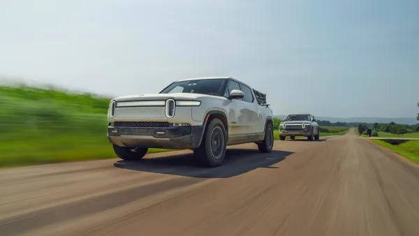 Two silver Rivian electric pick-up trucks driving down road with blurry grass on roadside