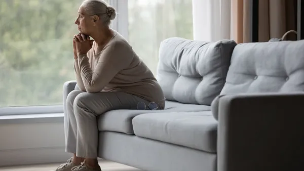Side view of older woman sitting on gray sofa gazing pensively out of wall-sized window.
