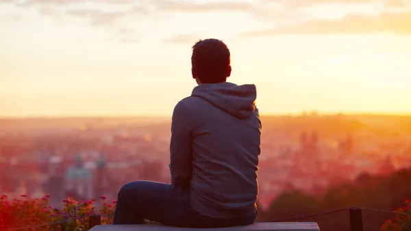 View from back of young man sitting on concrete bench overlooking blurred city at sunrise or sunset