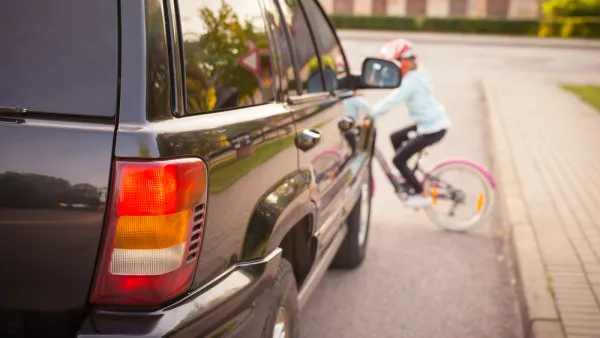 View of large black SUV from behind with blurred small child on bicycle wearing helmet riding in front of car